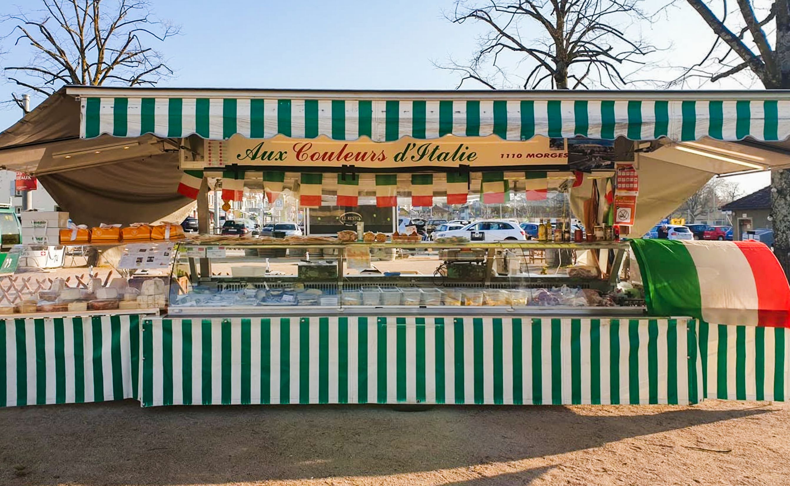 Le stand Aux Couleurs d’Italie sur un marché : auvent rayé vert et blanc, vitrine de charcuteries et fromages, fanions tricolores